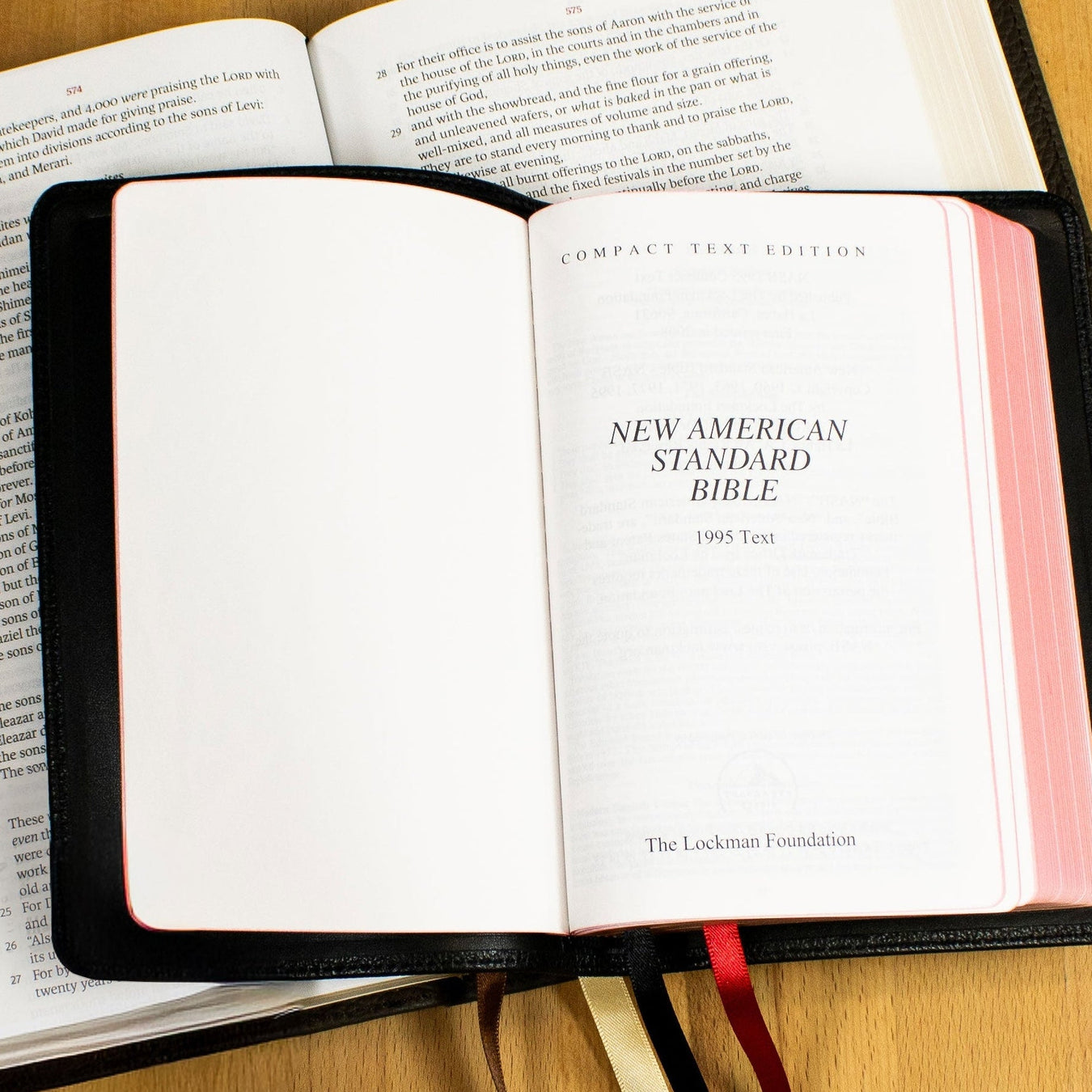 An open New American Standard Bible in a black hardcover lies on a light wood floor, with other open books partially visible underneath and ribbon bookmarks hanging from the pages.